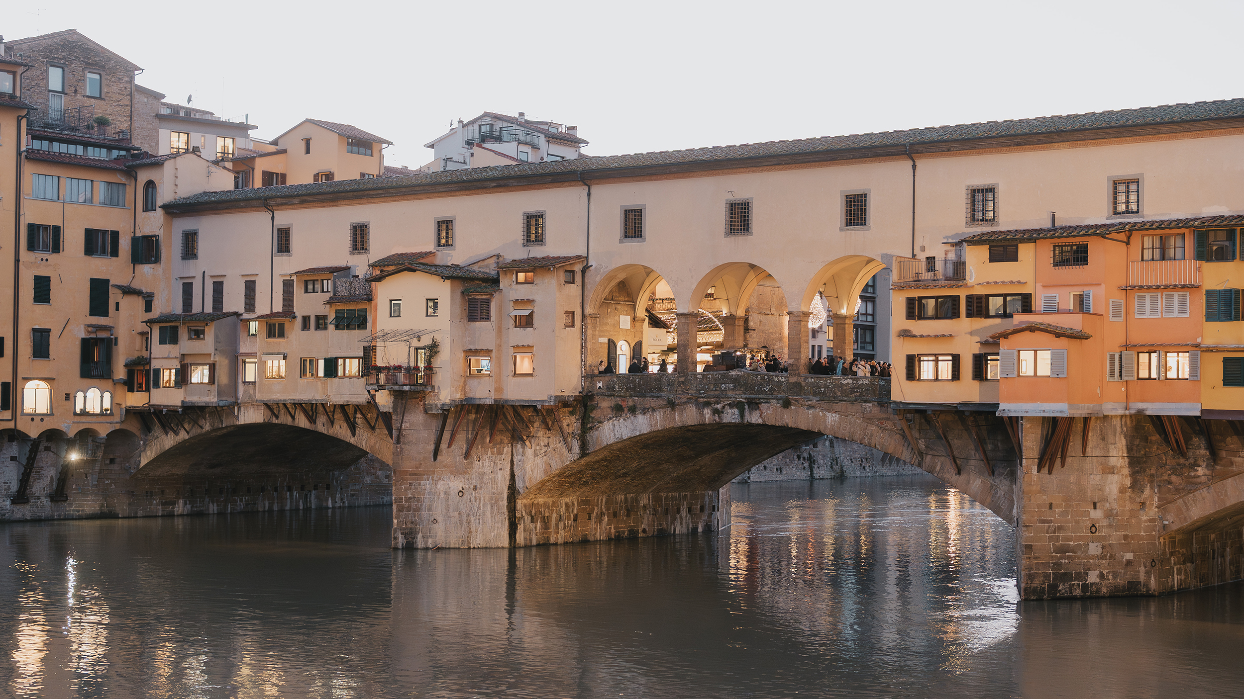 Ponte Vecchio at sunset over the Arno River in Florence, Tuscany, with warm lights reflecting on the water.