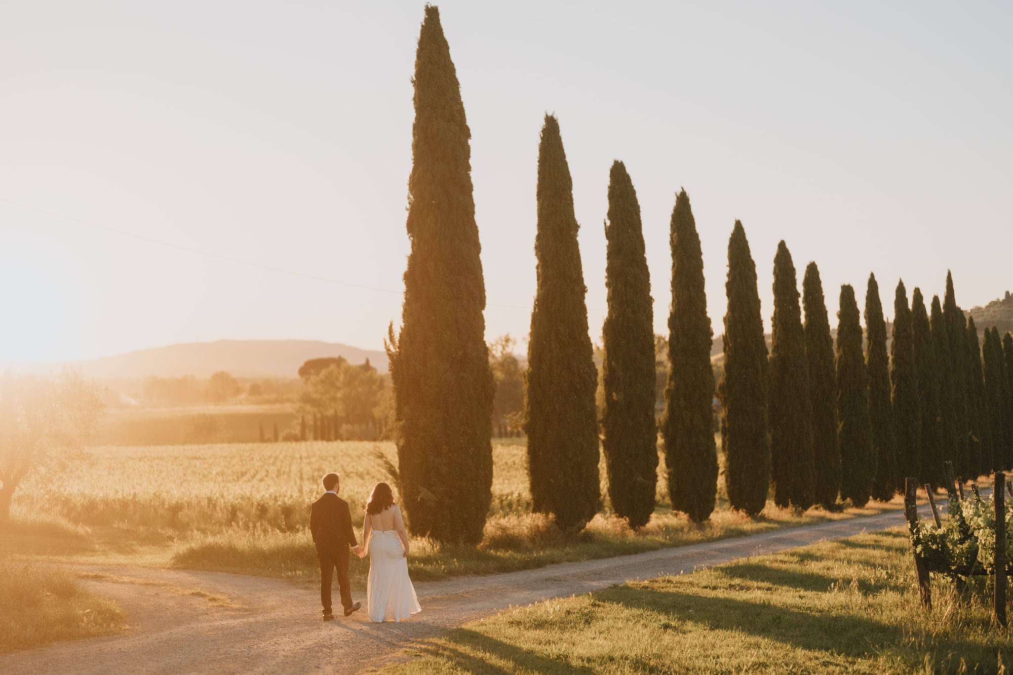 ride and groom walking away hand in hand along a cypress-lined gravel road at sunset, at La Sovana in Tuscany