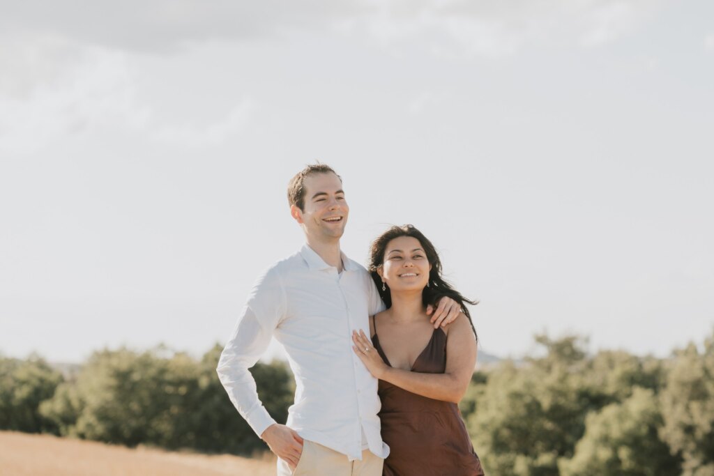 Engaged couple smiling arm in arm in the fields near the Chapel of Vitaleta