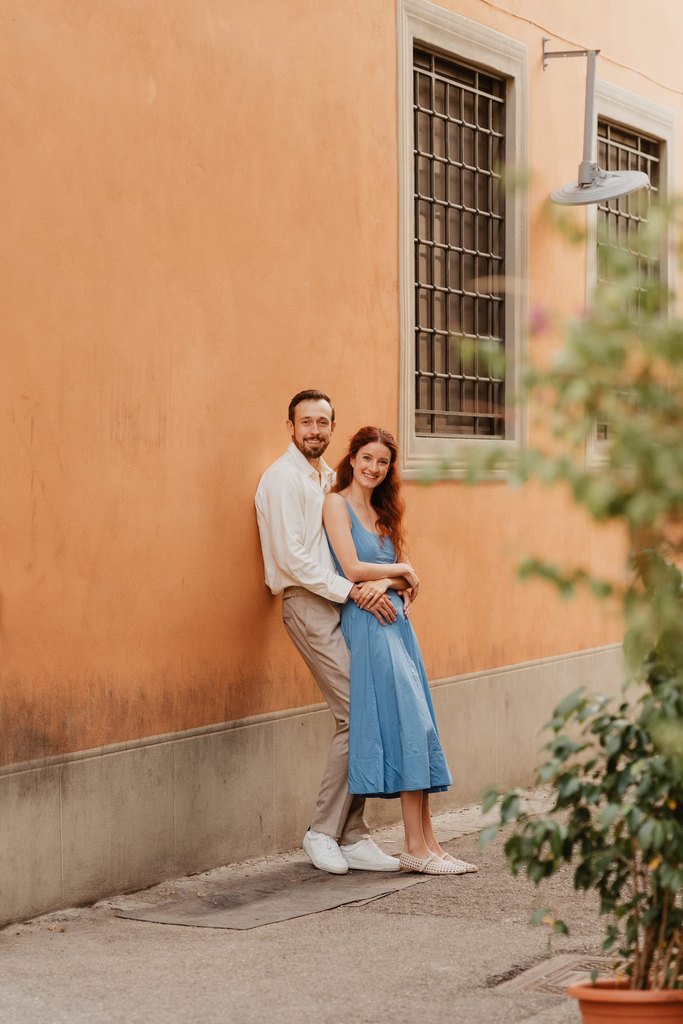 Couple leaning against an ochre wall in a Florence street during an engagement session, a barred window and a potted plant beside them