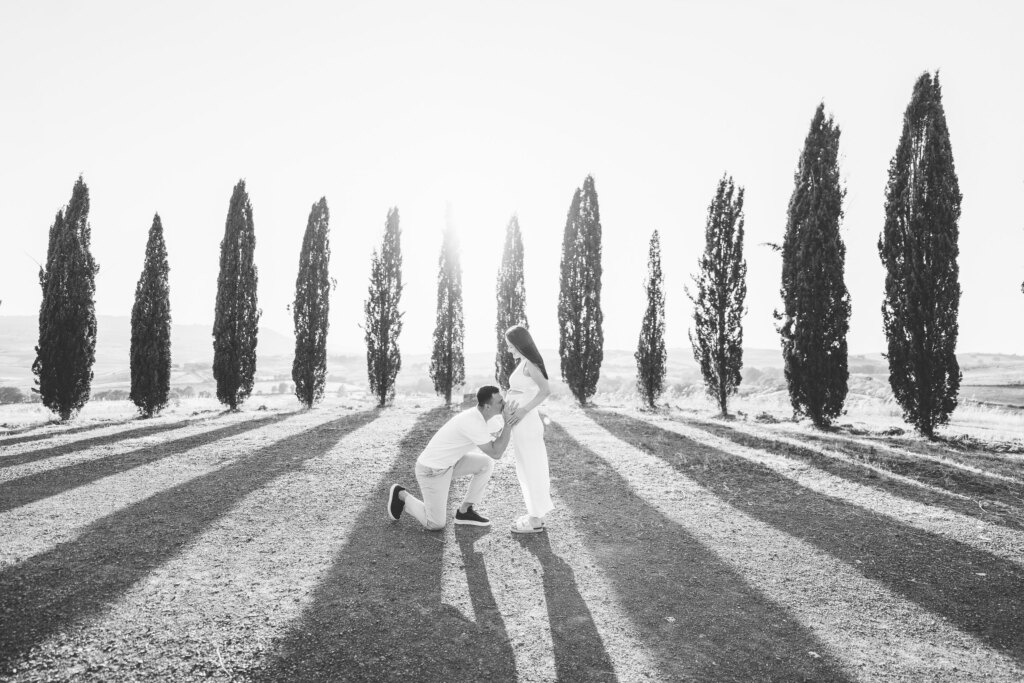 Maternity photoshoot Val d'Orcia: partner kneeling to kiss the baby bump among the cypresses near San Quirico d'Orcia