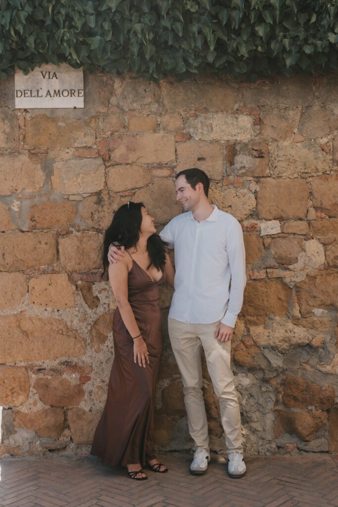 Engaged couple leaning on a stone wall under the Via dell'Amore street sign in Pienza