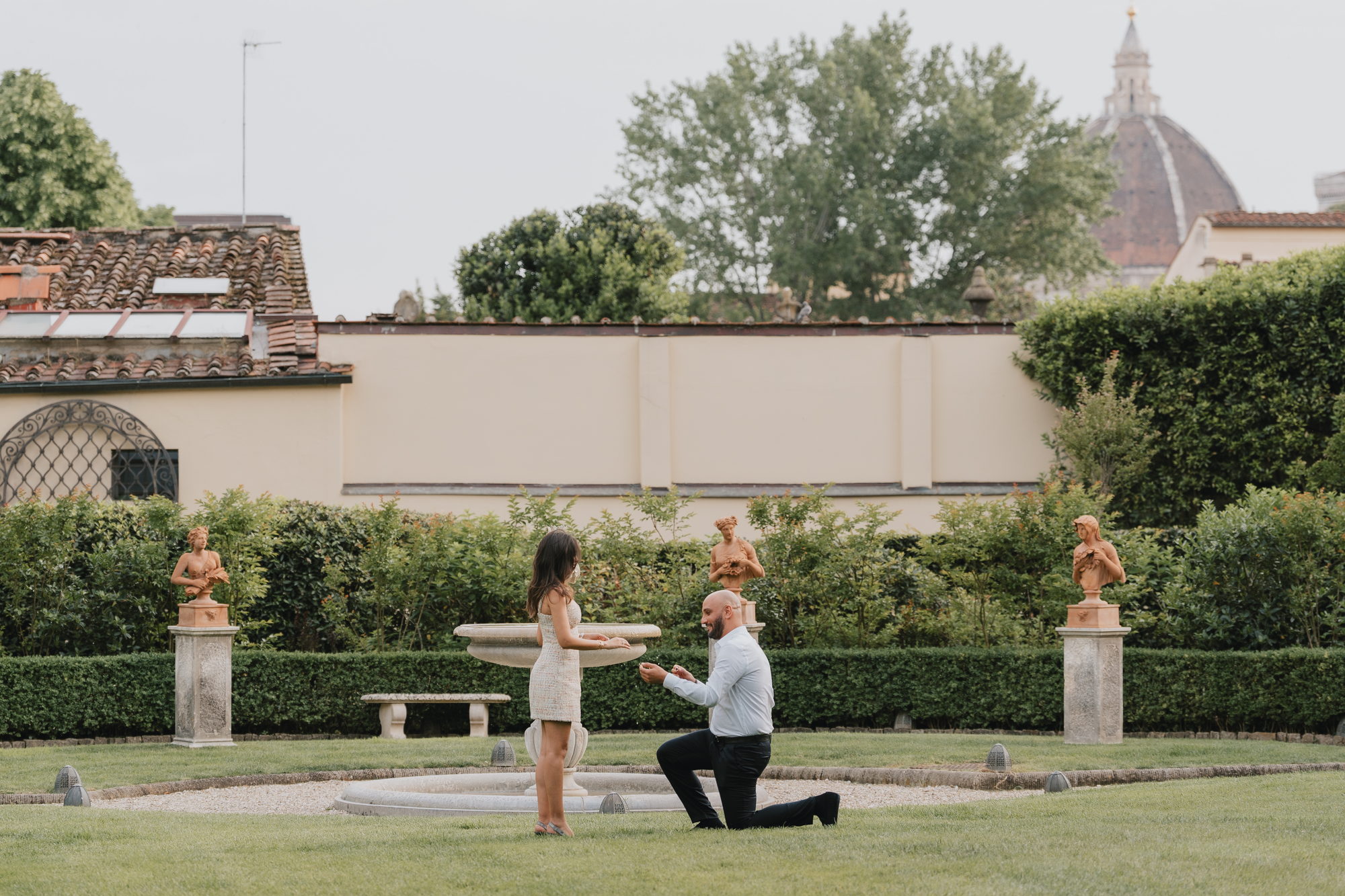 Man on one knee proposing with a ring in the garden of the Four Seasons in Florence, terracotta busts on pedestals and the Duomo dome behind them