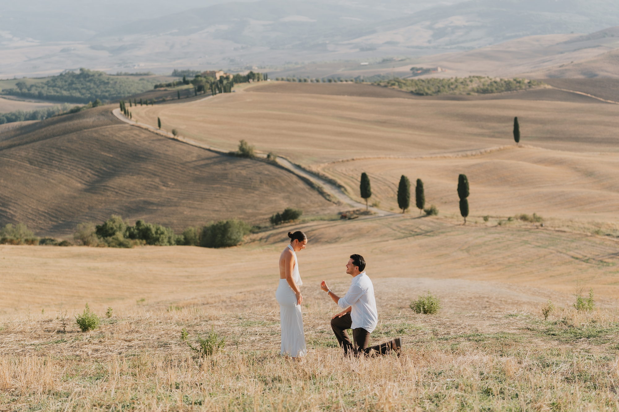 Man on one knee proposing with a ring in the Val d'Orcia hills near Pienza, cypress trees and a white road behind them
