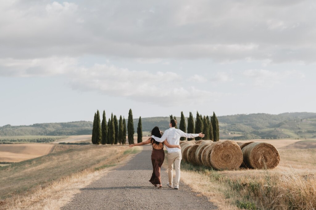 Couple walking arm in arm on the white road to the cypresses near San Quirico d'Orcia, round hay bales beside them