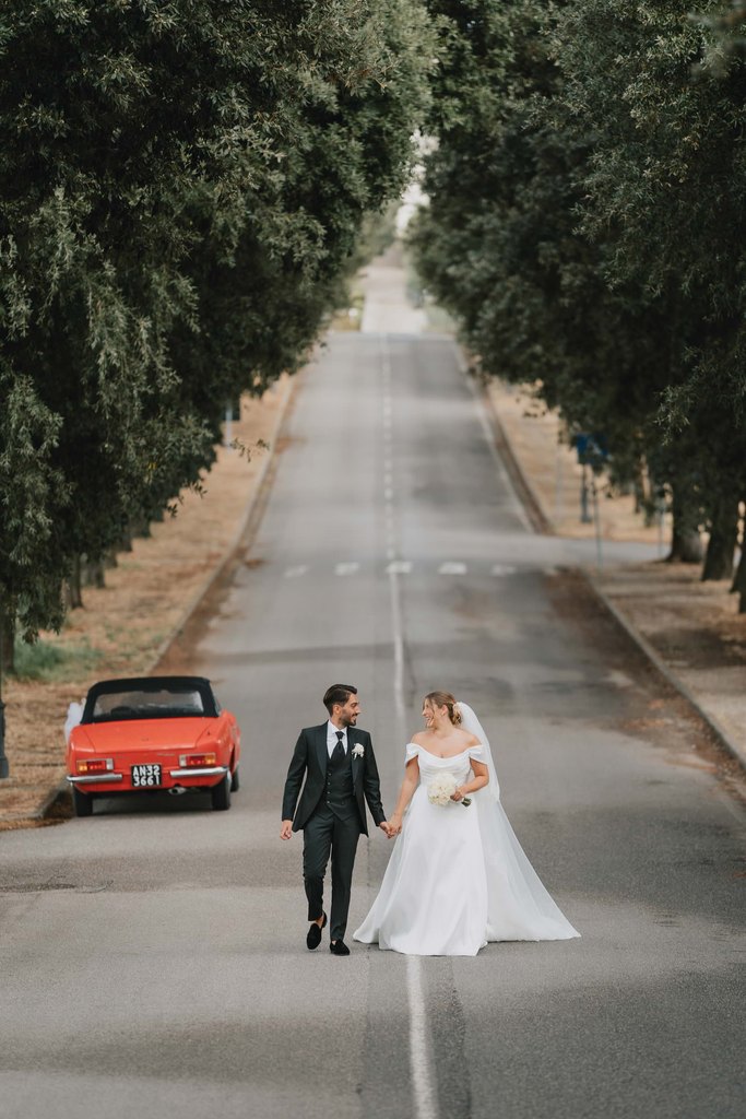 Bride and groom walking hand in hand down the tree-lined road at Artimino, near Florence, with a red vintage convertible parked behind them