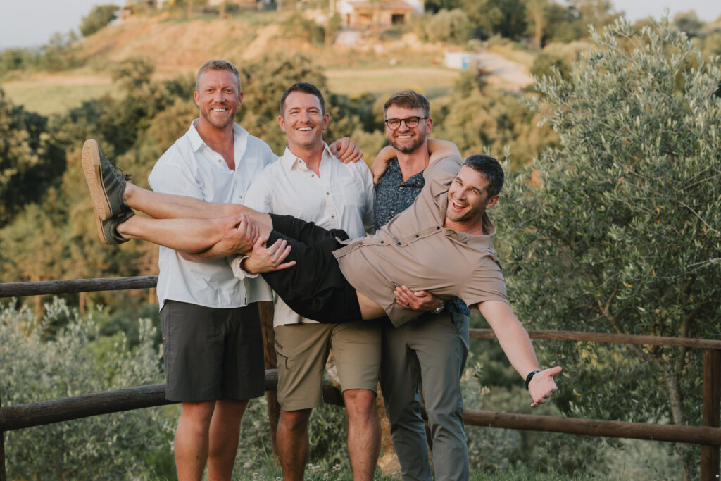 Tre uomini che sollevano in braccio il fratello su una terrazza affacciata sulle colline toscane