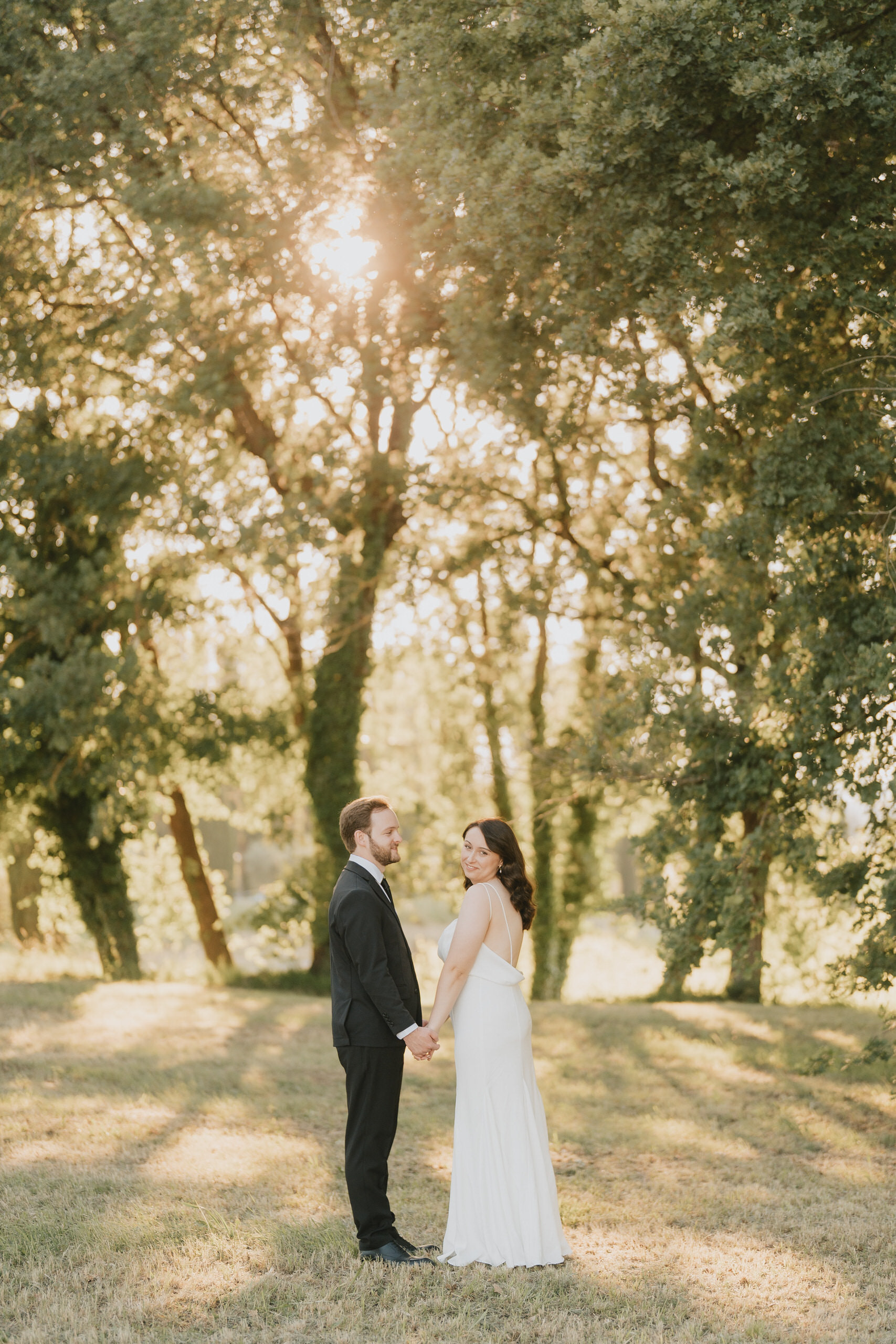Couple standing hand in hand under backlit trees at La Sovana