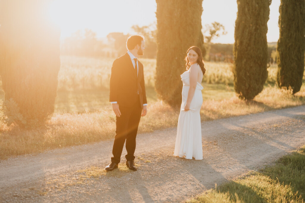 Bride and groom on a gravel road between cypresses in the late afternoon light