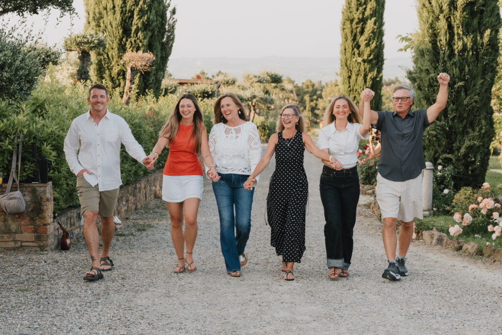 Famiglia che cammina mano nella mano verso la fotocamera lungo un vialetto di ghiaia tra i cipressi in Toscana