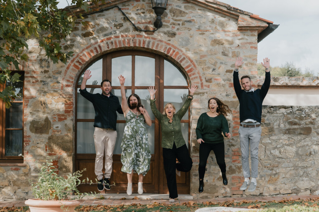 Famiglia di cinque persone che salta davanti al portone ad arco di un casale in pietra vicino a Siena