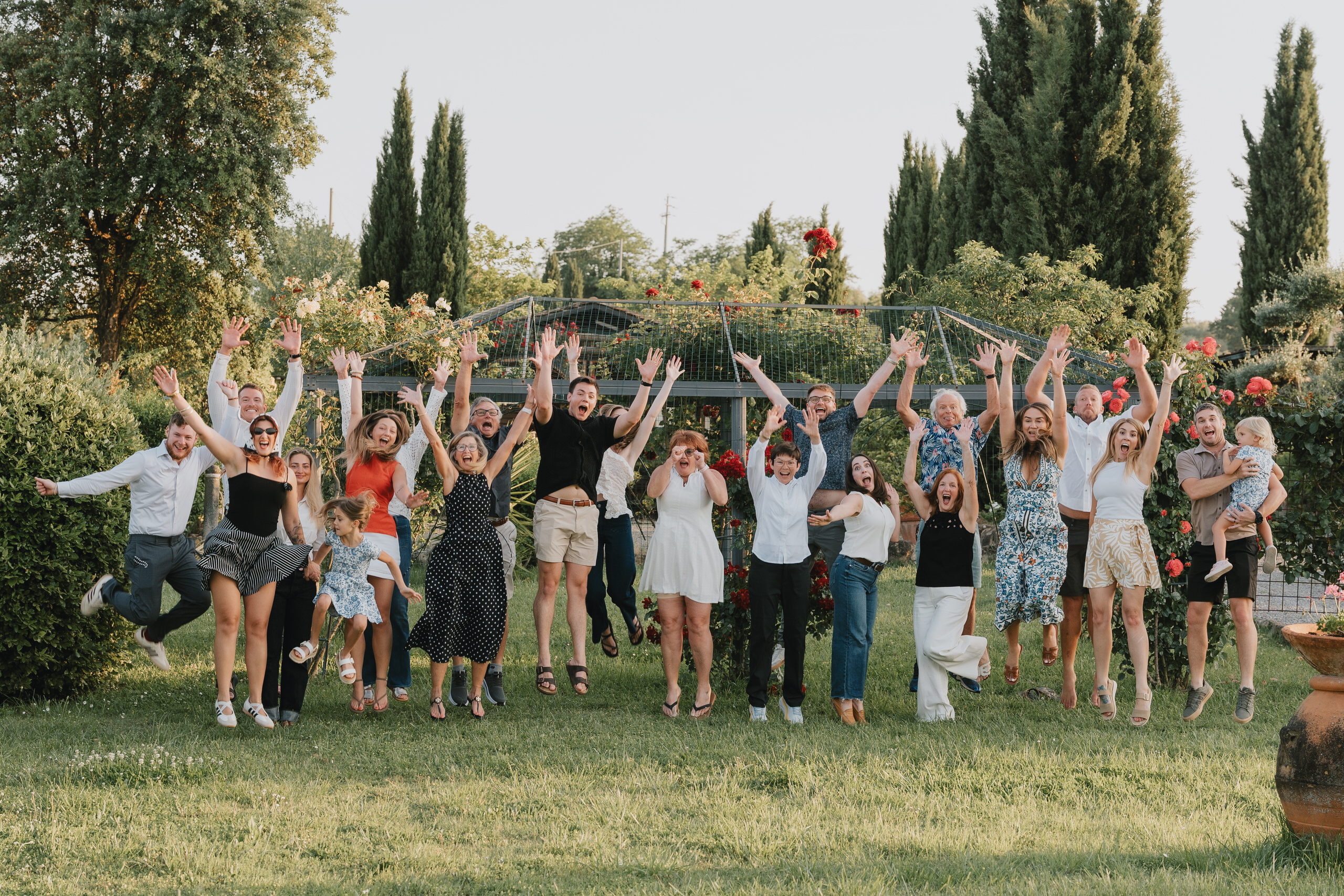 Grande famiglia riunita che salta insieme sul prato del giardino di una villa in Toscana, con cipressi e rose sullo sfondo