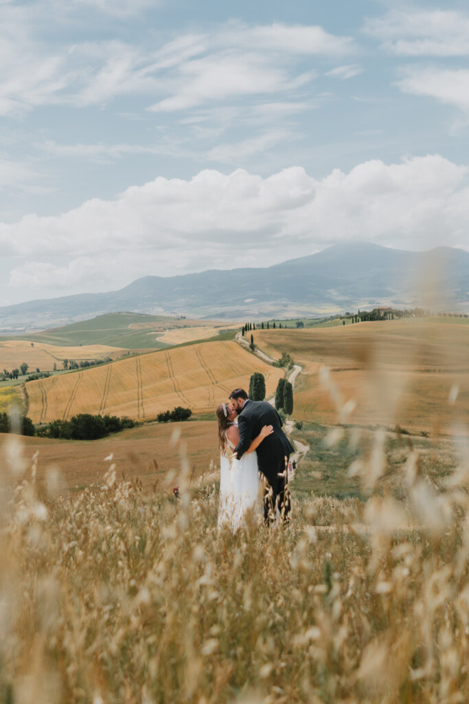 Bride and groom kissing in tall grass above a winding cypress road near Pienza
