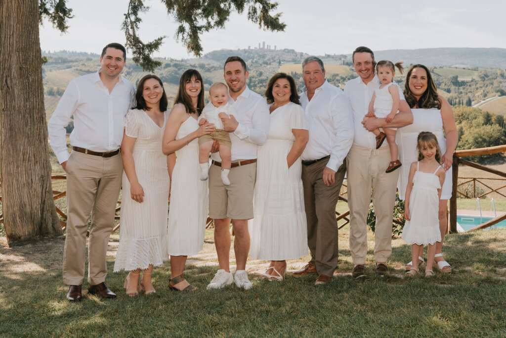Famiglia di tre generazioni vestita di bianco in posa sotto un albero, con le torri di San Gimignano sulle colline