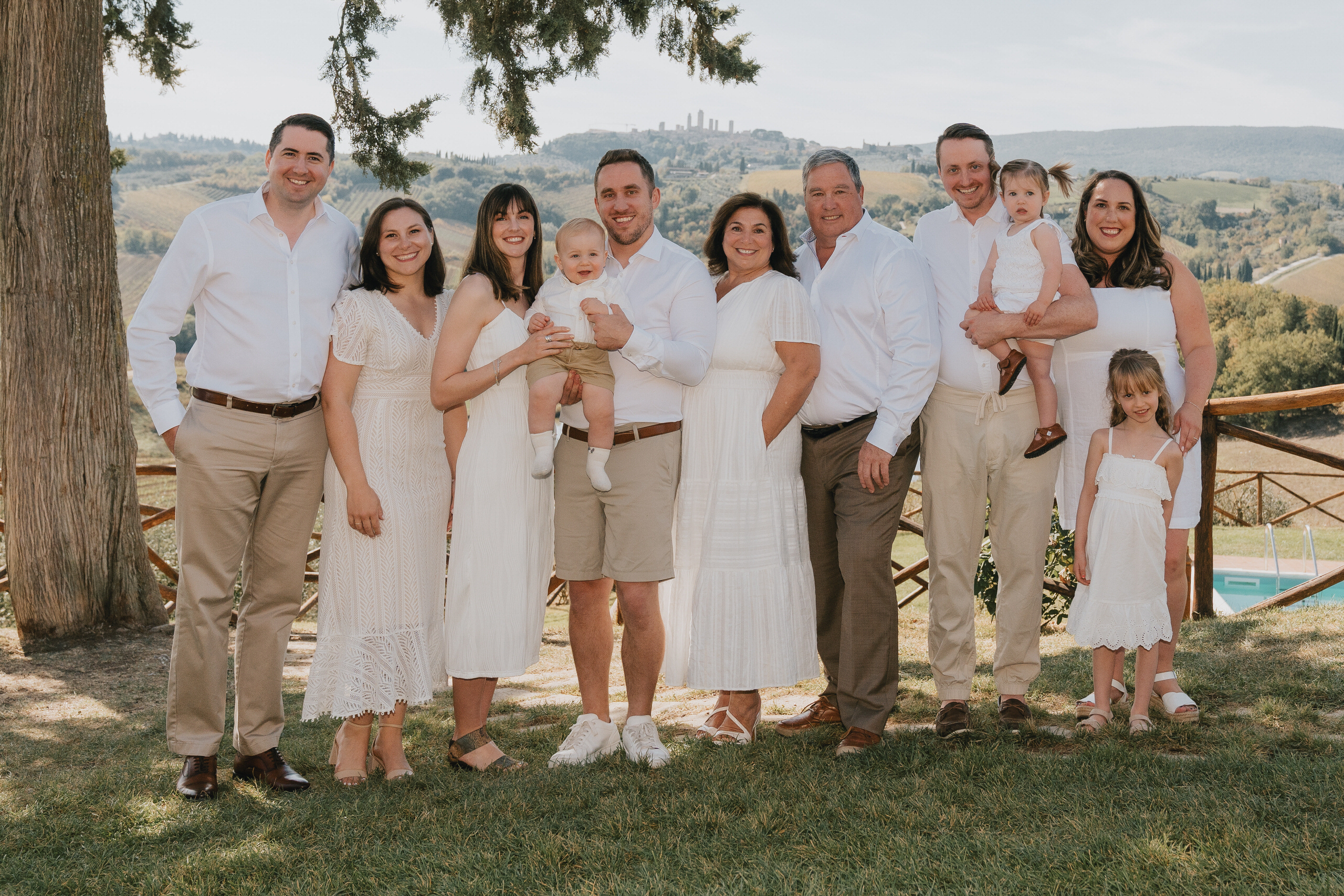 Famiglia di tre generazioni vestita di bianco in posa sotto un albero, con le torri di San Gimignano sulle colline