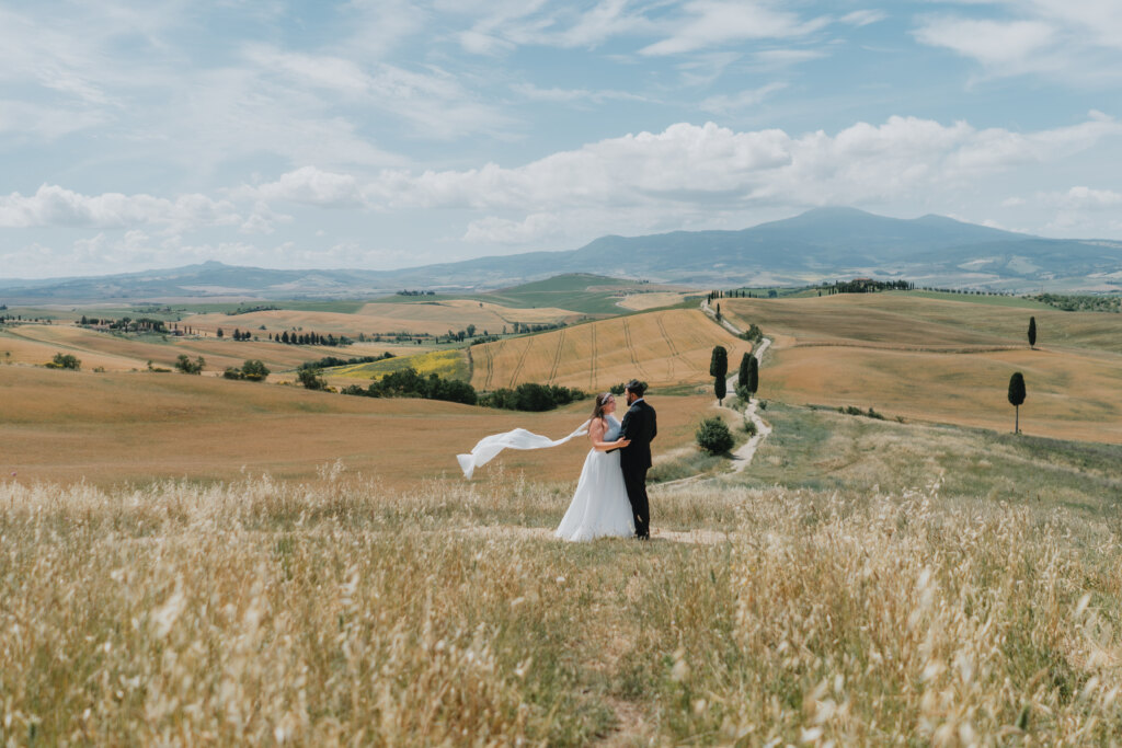 Pienza elopement: bride's veil lifted by the wind as the couple embraces above a cypress road in the Val d'Orcia