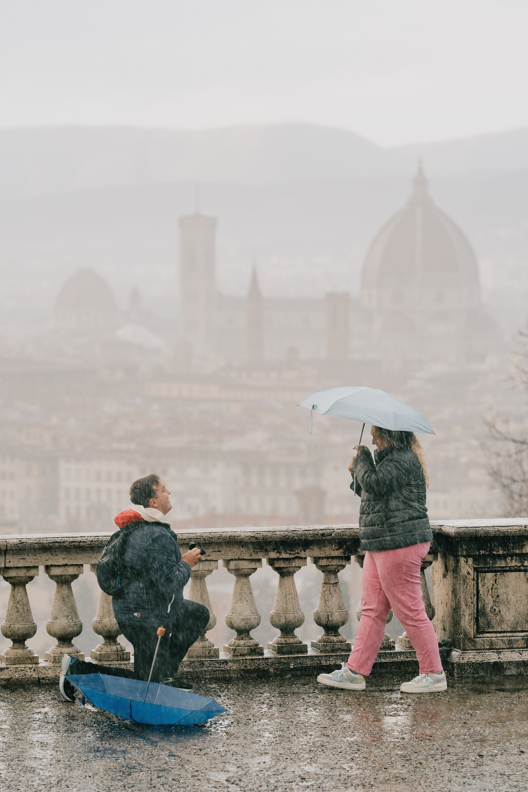 Rainy day proposal in Florence: he kneels on the wet terrace, she holds an umbrella