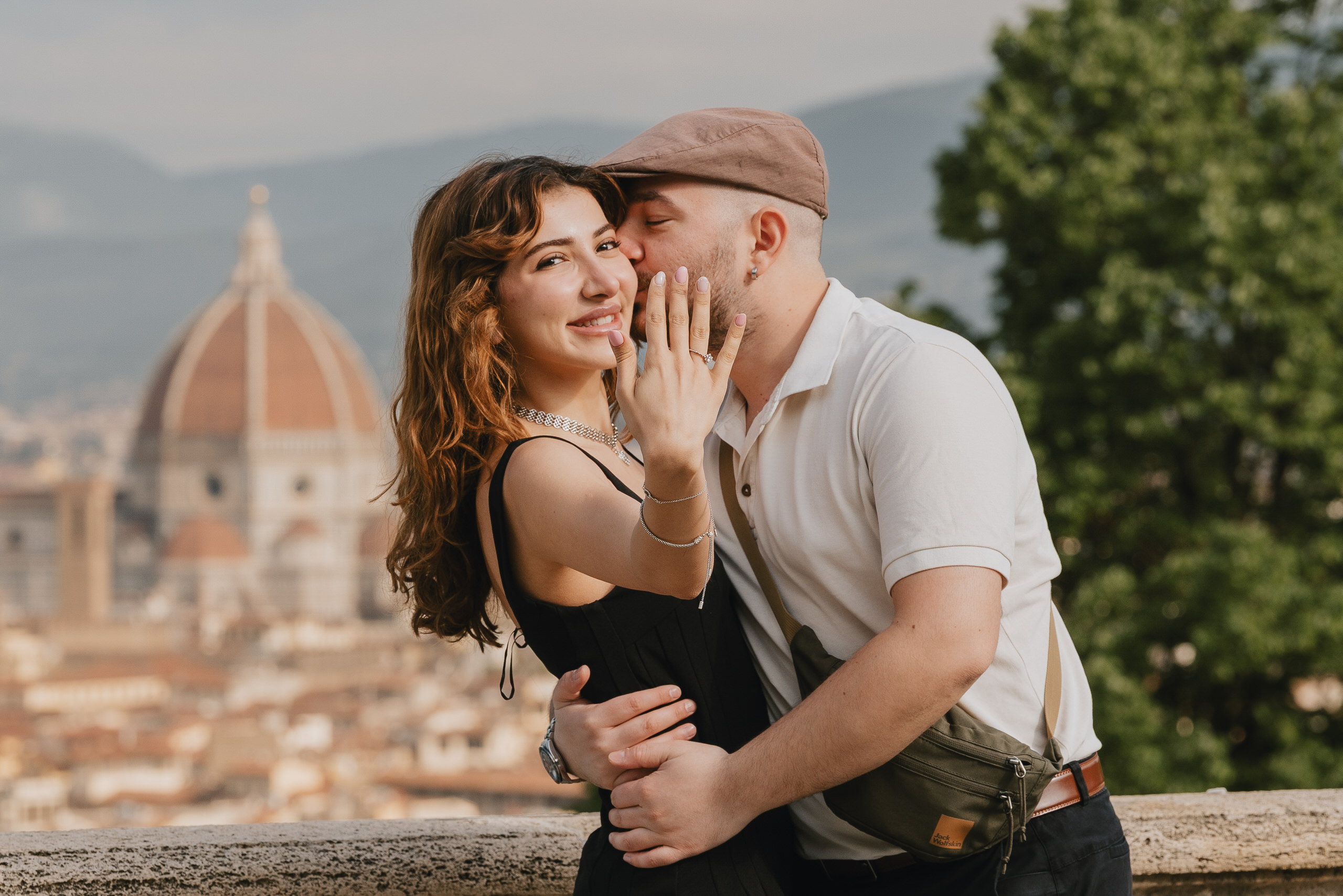 She shows the ring, he kisses her cheek, the Duomo of Florence behind