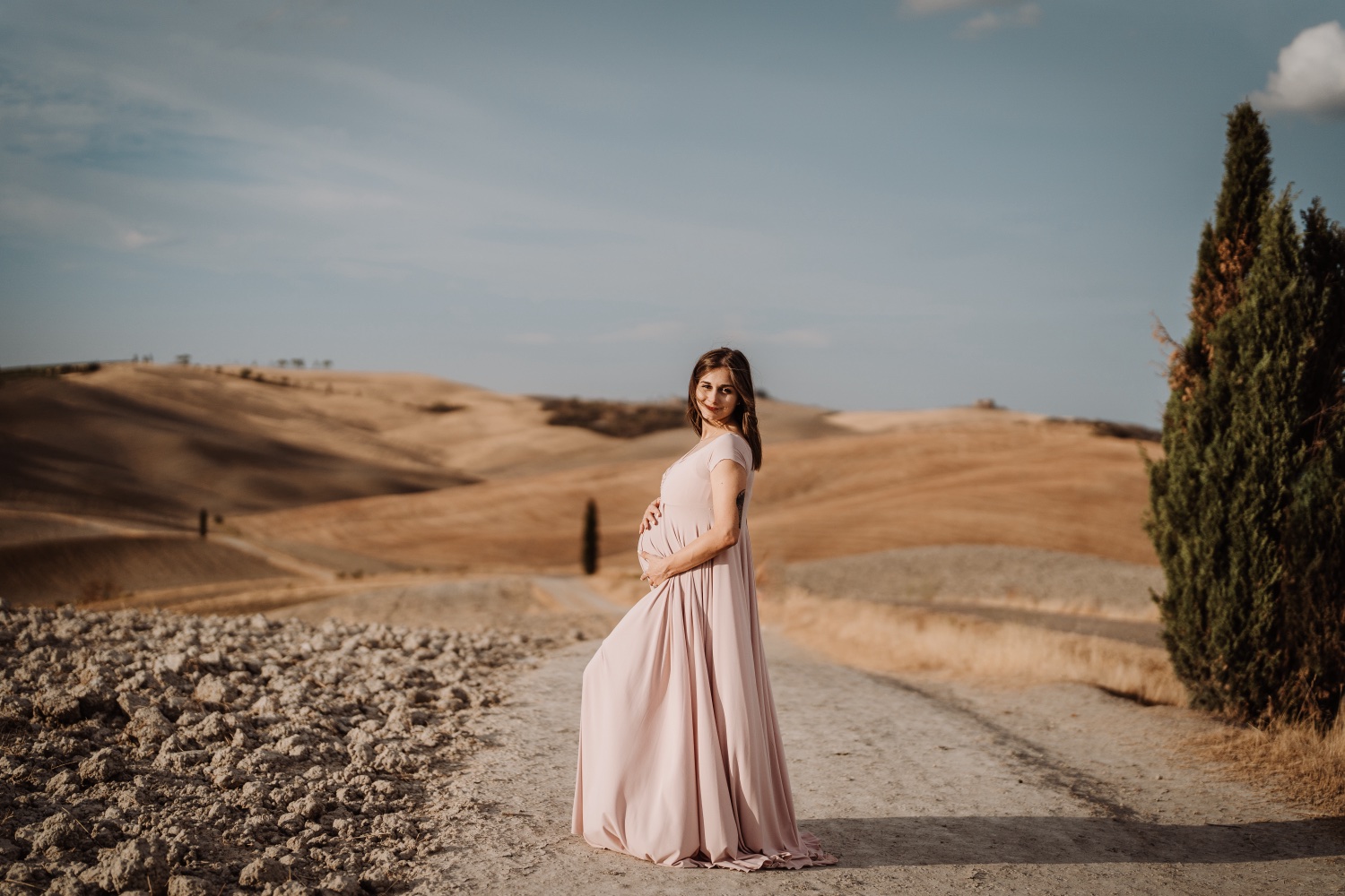 Futura mamma in abito rosa accanto a un cipresso, le colline dietro
